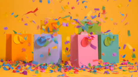 Studio-Shot-Of-Colourful-Birthday-Party-Gift-Bags-Against-Yellow-Background-With-Falling-Paper-Confetti